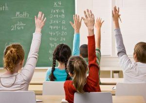 Students in classroom raising their hands to answer their teacher's question. Horizontal shot.