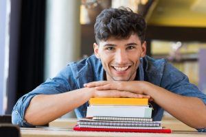 Closeup shot of young man leans on stack of books. Happy male student smiling and looking at camera. Lughing guy leaning on a pile of books and school notebooks.