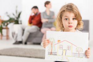 Upset little boy holding a drawing of a house, with his parents sitting angry on a couch in the blurry background